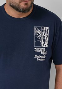 Behaarde man die een marineblauw T-shirt draagt met een witte grafiek van blote boomtakken en de tekst "Wayward Tracks Inc. & Scouts" en "Omarm het Onbekende."
