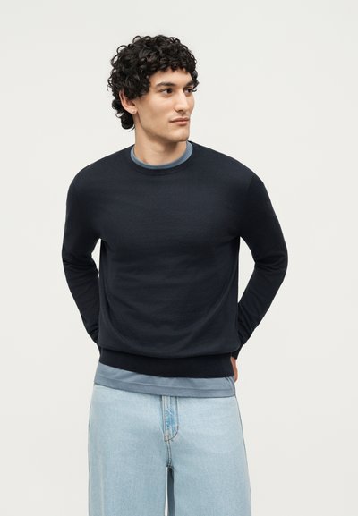 Νεαρός με κατσαρά μαλλιά που φοράει μαύρο πουλόβερ πάνω από μπλε πουκάμισο και ανοιχτό μπλε τζιν, στέκεται με τα χέρια πίσω από την πλάτη, κοιτώντας δεξιά.