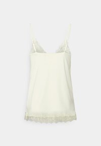 Krämfärgad linne med spetsdetaljer och justerbara spaghetti-band, med en vågig spetskant, mot en enkel vit bakgrund.