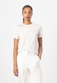 Light beige t-shirt features a subtle all-over pattern, short sleeves, and a classic crew neckline, paired with white trousers.