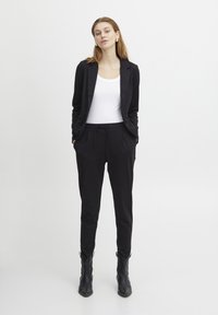 Donna in piedi con blazer nero, pantaloni neri, canotta bianca, stivaletti neri, mani in tasca, sfondo neutro.