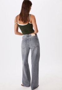 Femme portant un jean taille haute gris à jambes larges et un débardeur vert, debout de dos face à un fond blanc.