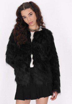 Zwarte faux fur jas met lange, gestructureerde mouwen en een ronde halslijn, gecombineerd met een witte top en een zwarte geplooide rok.