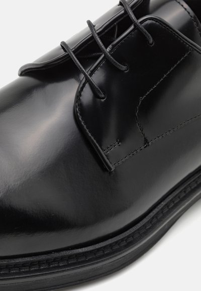 Scarpa da cerimonia in pelle nera con finitura lucida, caratterizzata da sei fori per i lacci, spessi lacci neri e una sottile cucitura lungo il bordo.