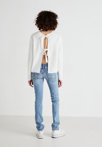 Vit ribbad tröja med öppen rygg som hålls på plats av en rosett, i kombination med ljusblå slim-fit jeans och vita sneakers. Slät struktur, avslappnad design.