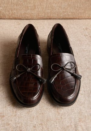 Hnedé kožené mokasíny s krokodílim vzorom, ozdobené mašľou s strapcami a plochou gumovou podrážkou.