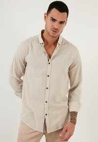 Camisa beige de botones con un cuello estructurado, mangas largas y una textura sutil. Presenta acentos de botones negros y un cuello interior en contraste.