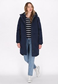 Marineblauwe geïsoleerde parka jas met een ritssluiting aan de voorkant, kap en twee zijzakken; gedragen over een zwart-wit gestreepte shirt en lichte jeans.