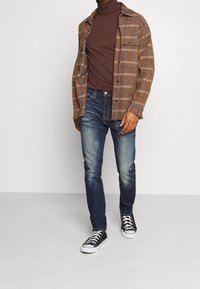 Hombre vestido con un suéter marrón de cuello alto, una camisa de cuadros marrón abierta, unos jeans azules deslavados y zapatillas negras de caña alta, de pie sobre un fondo claro liso.