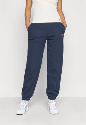 Navy jogger pants made of soft material with elastic cuffs and side pockets. Features pink logo embroidery on the thigh. Paired with white sneakers.