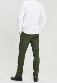 Pantalon vert en tissu texturé, coupe ajustée, associé à une chemise blanche à manches longues et des chaussures noires, vu de dos.