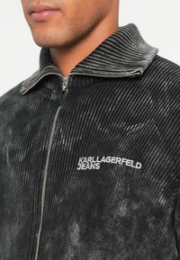 Fókuszált kép egy személyről, aki fekete és szürke bordázott zipzáros pulóvert visel, rajta a "KARL LAGERFELD JEANS" logóval a mellkasán.