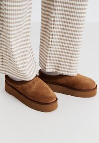 Brown suede slip-on shoes with a thick, textured sole, worn with white and tan striped wide-leg pants.
