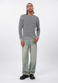 Uomo in piedi che indossa un maglione a righe grigie e nere, jeans larghi sbiaditi, scarpe nere e una collana con ciondolo a forma di croce su sfondo bianco.