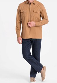 Brązowa koszula zapinana na guziki z dwoma kieszeniami na piersiach, w zestawieniu z ciemnymi jeansami i beżowymi trampkami. Minimalistyczny design i miękka tekstura.