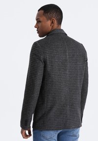 Grau karierter Blazer mit strukturiertem Design, texturiertem Stoff, Reverskragen und knöpfbaren Manschetten, getragen über blauen Jeans.