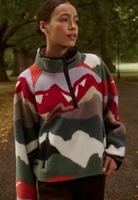 Pullover in pile con colletto alto, caratterizzato da un motivo astratto multicolore di montagne in rosso, verde e grigio. Include una tasca laterale con zip.
