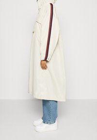 Béžový pláštěnka s kapucí, s červenými a námořnickými pruhy na rukávech, zapínáním na knoflíky a uvolněným střihem, nošená přes modré džíny a bílé boty.