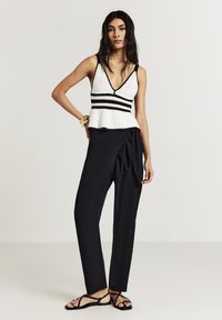 Femme debout portant un haut en crochet blanc sans manches avec des bordures noires, un pantalon noir taille haute à nouer, des sandales et des bracelets en or.