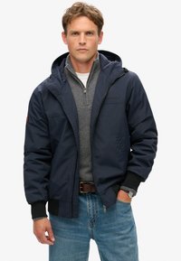 Mørkeblå vandtæt jakke med hætte, lynlås foran, elastiske manchetter og sidelommer, båret over en grå sweater og jeans.