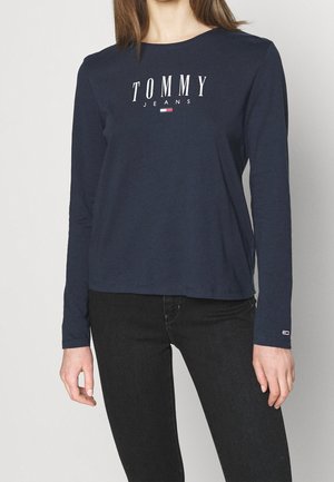Femme portant un t-shirt en bleu marine à manches longues Tommy Jeans avec un texte de logo sur la poitrine et un petit logo sur la manche gauche, assorti à un jean noir.