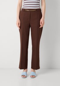 Pantaloni marroni su misura con un accento in catena dorata in vita; abbinati a sandali blu chiaro e una maglietta a righe.