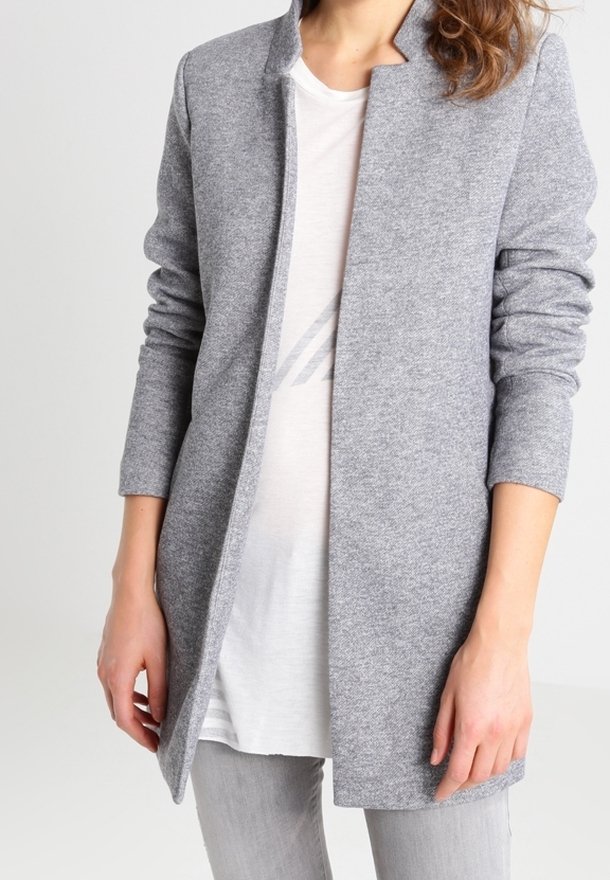 Femme portant un cardigan en maille gris clair ouvert sur un t-shirt blanc et un pantalon gris, debout avec les mains détendues.