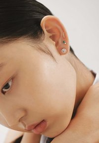 Tres piercings en la oreja: un pequeño stud con gema verde, un acento dorado y un stud redondo con cristal. Piel suave y cabello oscuro visibles en una vista en primer plano.