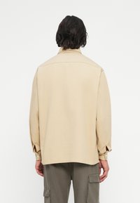 Persona con capelli corti scuri che indossa una camicia a maniche lunghe beige e pantaloni cargo grigi, vista di spalle su uno sfondo bianco.
