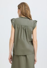 Femme aux cheveux châtain clair porte un haut sans manches vert olive avec des épaules à volants et une jupe assortie, vue de dos sur un fond uni.