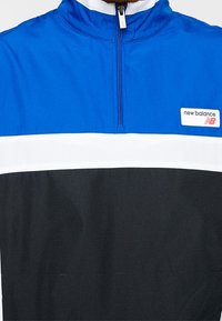 Blå och svart kvartslång zip-jacka med vit horisontell rand. Har en ståkrage och en logotag på vänster bröst. Texturerad tyg.