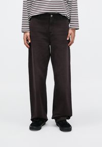 Person iført løse sorte bukser, sorte sko og en langærmet skjorte med sorte og hvide vandrette striber, stående mod en hvid baggrund.