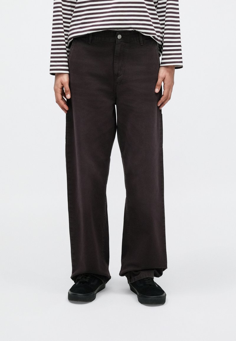 Persona vistiendo pantalones negros holgados, zapatos negros y una camisa de manga larga con rayas horizontales blancas y negras, de pie contra un fondo blanco.