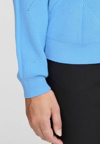 Maglione lavorato a maglia di colore azzurro chiaro con polsini a coste e motivi testurizzati, indossato sopra un indumento nero. Primo piano del maniche e della mano.