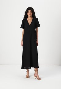 Black maxi dress with a V-neck, short puff sleeves, and lace trim. Smooth texture and flowy shape paired with strappy beige heels.