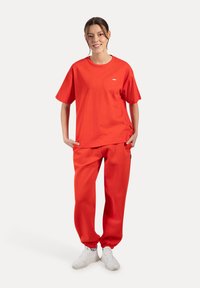 Giovane donna in piedi con le mani in tasca, che indossa una maglietta oversize rossa brillante abbinata a pantaloni della tuta rossi e sneaker bianche.