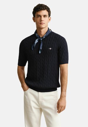 Man draagt een marineblauwe kabelgebreide trui met korte mouwen, een lichte sjaal om de hals geknoopt en gebroken-witte broek, staat met één hand in de broekzak.