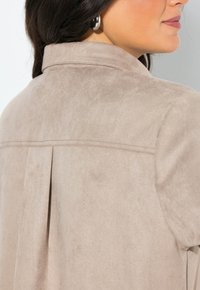 Helle taupe-farbene Kunstwildlederjacke mit einer strukturierten Oberfläche, einem Kragen und einer Rückenfalte. Mit schlichten Nähten und runden metallischen Ohrringen.