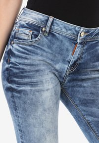 Blauwe denim jeans met een vervaagde afwerking, voorzien van oranje en groene stikselaccenten, vijf zakken en een ritssluiting met een knoop.