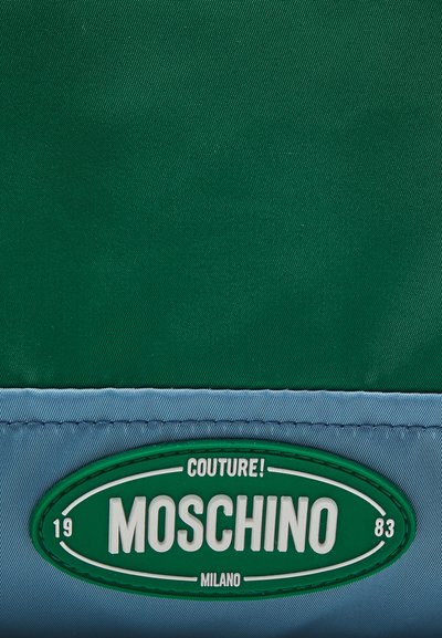 Green and blue nylon fabric with a smooth texture. Features a round Moschino logo patch in green and white stitched at the bottom.