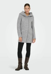 Manteau texturé gris avec une grande capuche, des boutons à l'avant et des manches longues. Associé à des leggings noirs et des bottes marron jusqu'aux genoux.