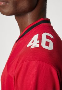 Sarkanais krekls ar teksturētu audumu, ar V veida kakla izgriezumu un melnu apdari. Plecā atrodas numurs "46" baltā krāsā ar "EST." detaļu.