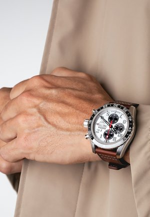 Handgelenk mit silberner Chronograph-Uhr, weißem Zifferblatt, schwarzen Unterzifferblättern, rotem Sekundenzeiger, drehbarer Lünette und braunem Lederarmband über einem beigen Ärmel.