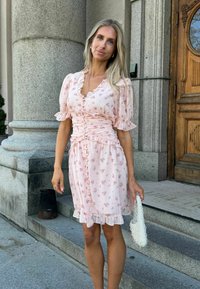 Femme aux longs cheveux blonds porte une robe à fleurs rose et tient une pochette blanche perlée, debout sur des marches en pierre près d'une porte en bois.