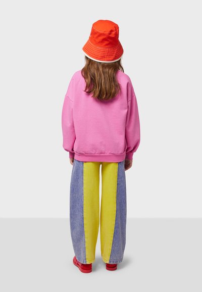 Bright pink oversized sweatshirt, yellow and blue striped wide-leg jeans, red bucket hat, and red shoes. Soft materials and casual fit.