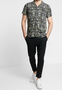 Man med kortärmad mönstrad skjorta med knappar, svarta slim-fit byxor, vita sneakers, händerna i fickorna, stående mot en enfärgad bakgrund.