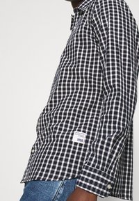 Camicia a quadri in blu navy e bianco, con colletto botton down, maniche lunghe e un'etichetta sul lato inferiore con l'anno "1990".