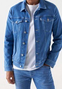 Muž v modré denimové bundě přes bílé tričko a odpovídajících modrých denimových džínsech, s jednou rukou v kapse a neutrálním pozadím.