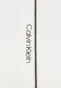 Bílá tkanina s ribbed texturou, se výraznou černou vertikální linií a logem "Calvin Klein" v černém písmu na boku.
