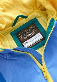Veste bleue à lignes jaunes avec une fermeture éclair ouverte montrant une étiquette portant l'inscription "Next Originals 1982" et un badge nominatif vierge.